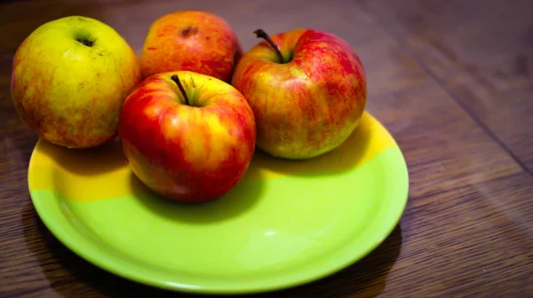 Four Ripe Red And Green Apples Are On The Green Round Plate On A Wooden Table.