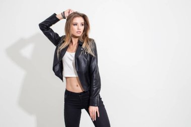 Deri ceketli ve siyah pantolonlu beyaz arka planda poz veren bir kız. Moda çekimi.