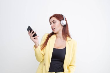 Sarı elbiseli kızıl saçlı bir kız büyük kulaklıklarla müzik dinliyor ve telefonda yazıyor.