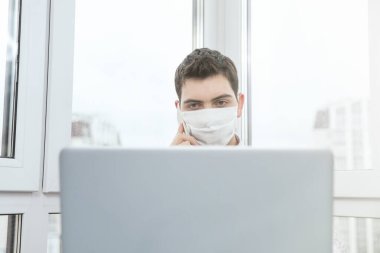 Koruyucu maskeli bir adam telefonda konuşuyor ve virüs salgını sırasında evden bilgisayar başında çalışıyor. Virüs enfeksiyonu sırasında karantina