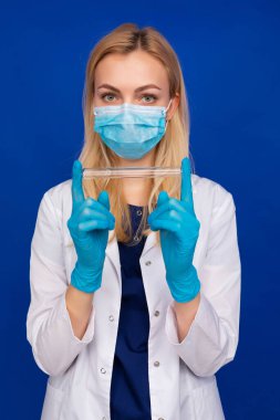 Tıbbi maskeli ve eldivenli bir kız doktor mavi arka planda test tüpü tutuyor.