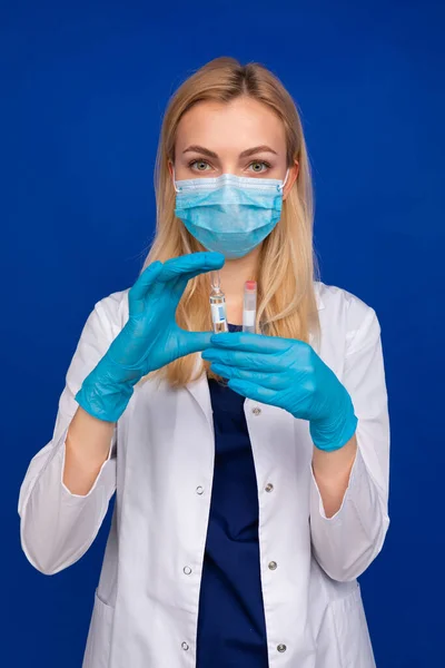 Tıbbi maskeli ve eldivenli bir kız doktor mavi arka planda ilaç ve test tüpü bulunan bir ampul tutuyor.