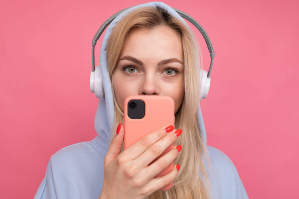 A girl in a fashionable hoodie listens to music from her phone in a good mood on a pink background