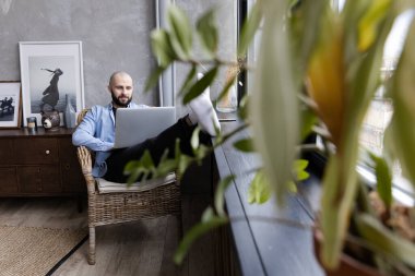 Mavi gömlekli adam evde, pencerenin yanında bir dizüstü bilgisayar üzerinde çalışıyor. Uzaktan çalışma kavramı.