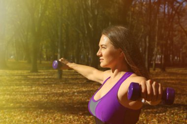 Parlak eşofmanlı bir kız parkta dambıllarla egzersiz yapıyor. Ayarlanmış