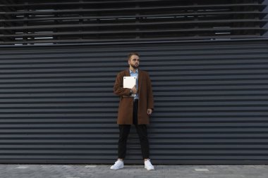 Paltolu ve dizüstü bilgisayarı olan metal bir duvara poz veren genç bir adam.