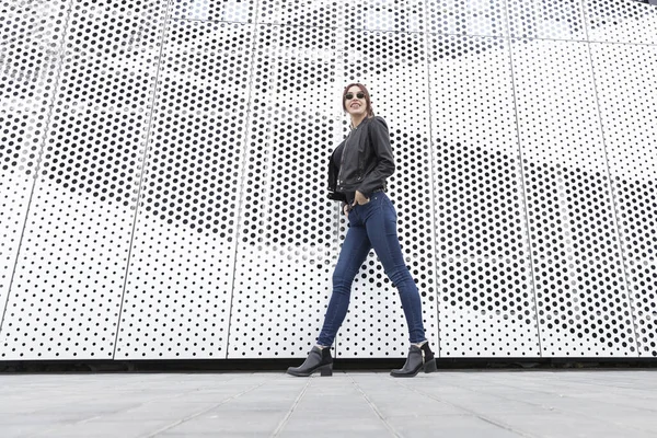 The red-haired girl in jeans and a leather jacket posing against the backdrop of a metal wall