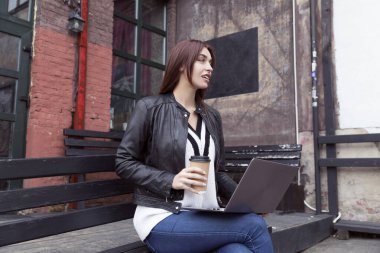 Siyah deri ceketli kızıl saçlı kız kahve içiyor ve laptopta çalışıyor. Sokakta bir bankta oturuyor.