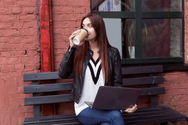 Siyah deri ceketli kızıl saçlı kız kahve içiyor ve laptopta çalışıyor. Sokakta bir bankta oturuyor.