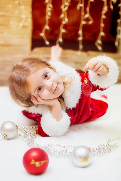 Menina vestida de Papai Noel com bolas de Natal em casa junto à lareira. No Natal. Feriados . — Fotografia de Stock