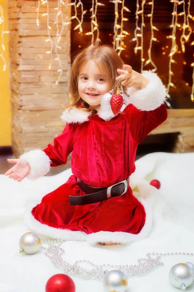 Küçük kız Noel Baba olarak şöminenin yanında evde Noel topları ile giyinmiş. Noel. Tatil. Telifsiz Stok Imajlar
