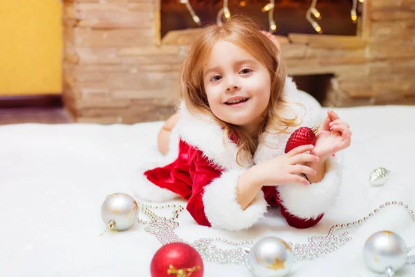 小女孩打扮成圣诞老人与圣诞球在家里壁炉旁。圣诞节。假期. — 图库照片