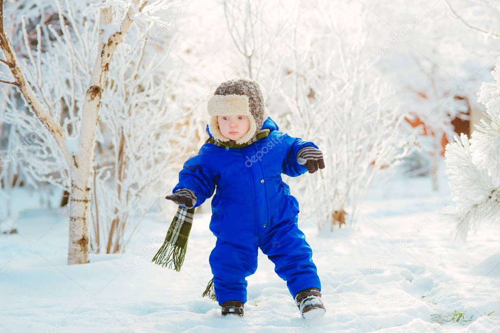 Child in the park with snow in winter. A little boy in blue overalls and a knitted hat, catches snowflakes in a winter park for Christmas. Children play in the snow-covered forest
