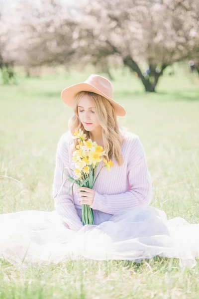 一个年轻漂亮的金发女孩在帽子和一条郁郁葱葱的长裙 春天花园里的水仙花花束 春天里捧着水仙花的年轻女士 — 图库照片