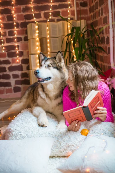 家で金髪の巻き毛の可愛い10代の女の子は大きな口の中の犬と枕の上にあり 本を読んでいます 人と犬 秋の気分 — ストック写真