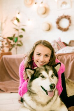 Cute teenage girl with blond curly hair at home, on the bed in the loft with a big malamute dog. People and Dogs. clipart