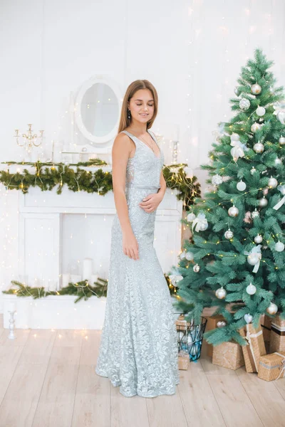 Attractive young girl with blond hair and evening make-up in an evening dress in a floor dress near a Christmas tree and a fireplace in a bright studio. New Year 2020