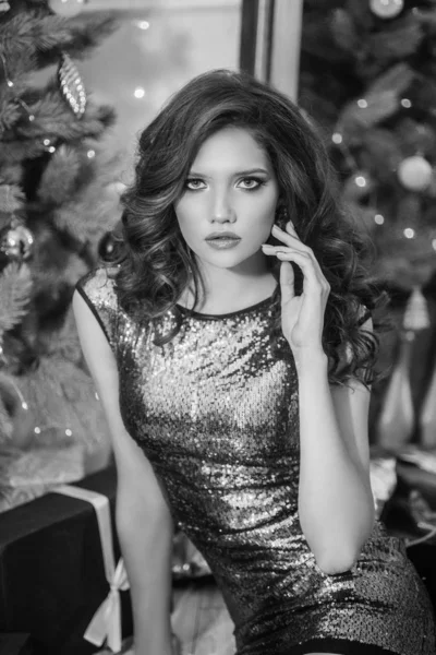 Attractive young girl with evening hairstyle and evening make-up in an evening sparkling dress near a Christmas tree and fireplace in a bright studio. New Year 2020