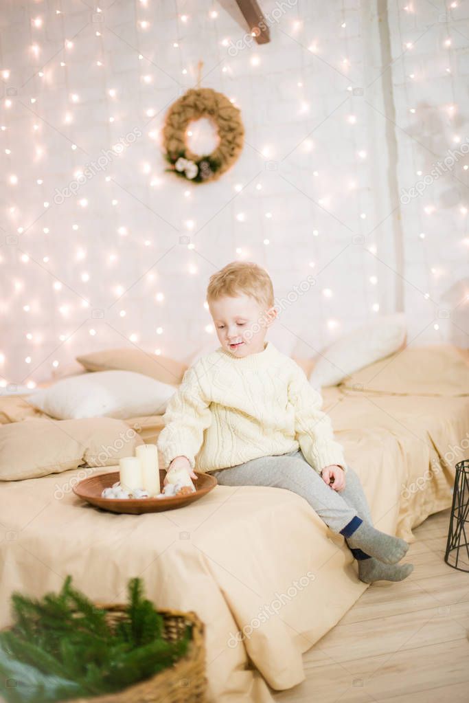 Cute little boy with blond hair in a light dark knitted sweater at home on the bed in a bright bedroom in a loft style decorated with Christmas garlands and needles. Christmas mood.