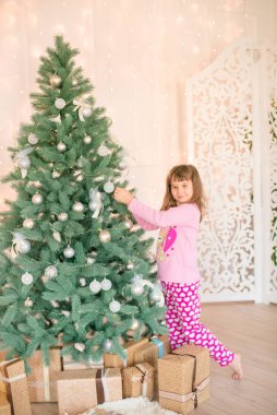 Noel ağacının yanında, üzerinde hediyeler, çelenkler, iğneler ve mumlarla süslenmiş parlak bir şömine olan pijamalı küçük şirin bir kız. Mutlu noeller. Noel havası
