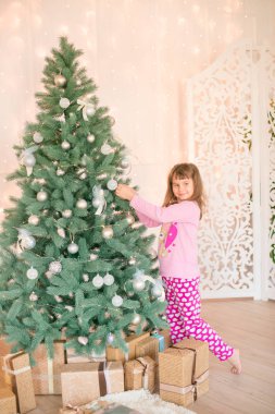 Noel ağacının yanında, üzerinde hediyeler, çelenkler, iğneler ve mumlarla süslenmiş parlak bir şömine olan pijamalı küçük şirin bir kız. Mutlu noeller. Noel havası