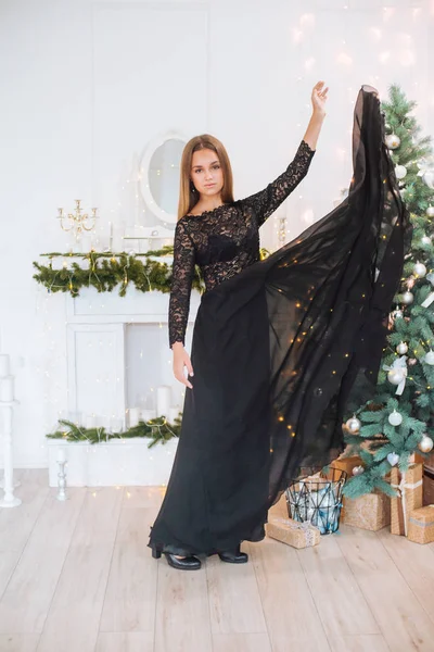 Attractive young girl with blond hair and evening make-up in an evening dress in a floor dress near a Christmas tree and a fireplace in a bright studio. New Year 2020