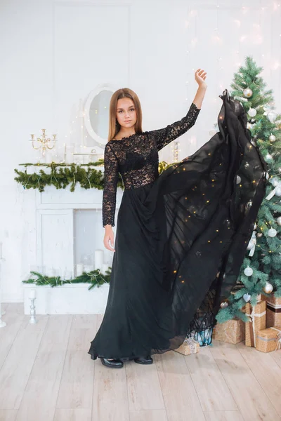 Attractive young girl with blond hair and evening make-up in an evening dress in a floor dress near a Christmas tree and a fireplace in a bright studio. New Year 2020