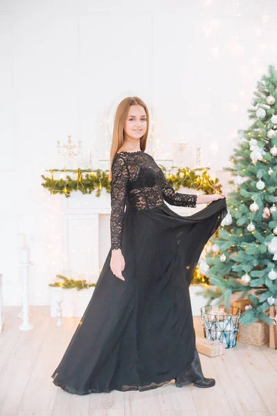 Attractive young girl with blond hair and evening make-up in an evening dress in a floor dress near a Christmas tree and a fireplace in a bright studio. New Year 2020