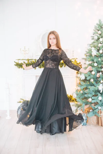 Attractive young girl with blond hair and evening make-up in an evening dress in a floor dress near a Christmas tree and a fireplace in a bright studio. New Year 2020