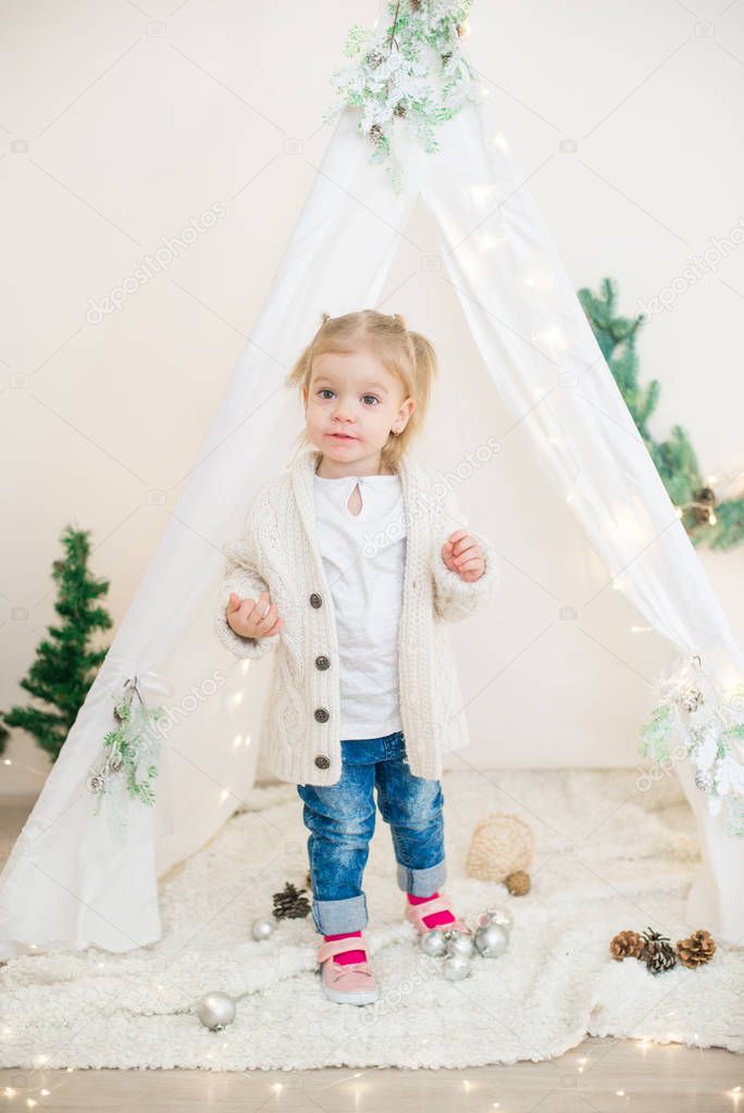 Little cute girl in a white warm knitted cardigan near a children's lodge decorated with a garland and needles, toys and tangerines. Christmas mood