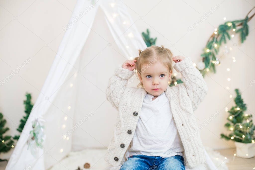 Little cute girl in a white warm knitted cardigan near a children's lodge decorated with a garland and needles, toys and tangerines. Christmas mood