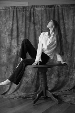 Beyaz tişörtlü, siyah pantolonlu, uzun saçlı ve tekstil arka planında stüdyoda poz veren güzel bir manken. Güzellik ve moda.