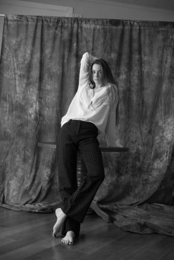 Beyaz tişörtlü, siyah pantolonlu, uzun saçlı ve tekstil arka planında stüdyoda poz veren güzel bir manken. Güzellik ve moda.