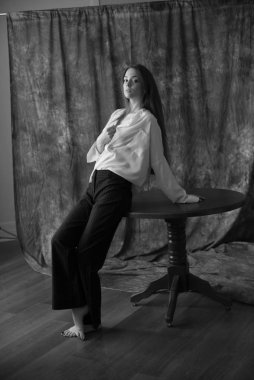 Beyaz tişörtlü, siyah pantolonlu, uzun saçlı ve tekstil arka planında stüdyoda poz veren güzel bir manken. Güzellik ve moda.