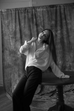 Beyaz tişörtlü, siyah pantolonlu, uzun saçlı ve tekstil arka planında stüdyoda poz veren güzel bir manken. Güzellik ve moda.