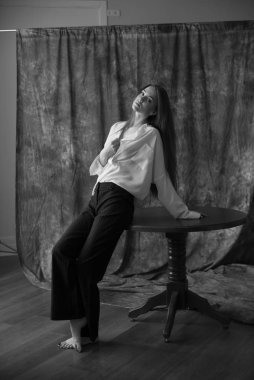 Beyaz tişörtlü, siyah pantolonlu, uzun saçlı ve tekstil arka planında stüdyoda poz veren güzel bir manken. Güzellik ve moda.