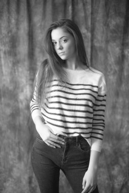 Çizgili kazaklı ve dar kotlu koyu renk uzun saçlı güzel bir manken stüdyoda tekstil arka planında poz veriyor. Güzellik ve moda.