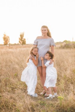 Sarışın, genç ve güzel hamile bir anne ve beyaz elbiseli iki kız gün batımında bir tarlada. Mutlu hamilelikler. Büyük aile. Aile ve Çocuklar