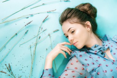 Siyah saçlı, klasik saç stili olan ve stüdyonun mavi arka planında lavanta çiçekleri olan açık renkli bir elbise giyen güzel bir genç kız manken. Güzellik ve moda.