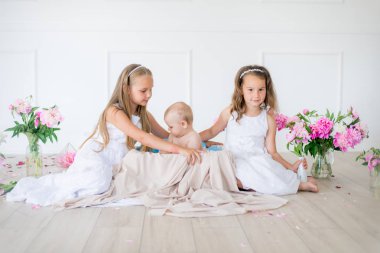 Beyaz elbiseli şirin küçük kız kardeşler ve pembe ve beyaz şakayıklarla süslenmiş açık bahar stüdyosunda bir erkek kardeş.