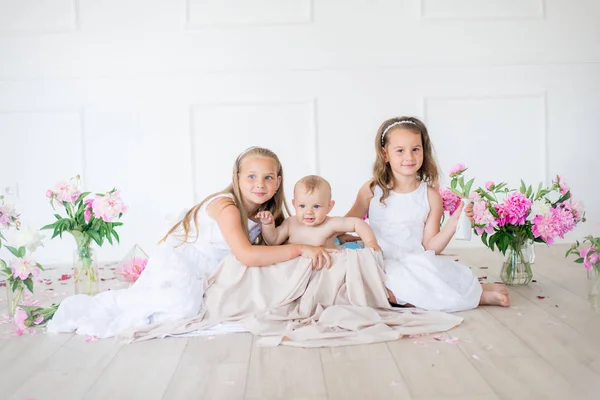 可愛いです小さな姉妹とともにブロンド髪で白いドレスと弟で光春のスタジオ装飾されたピンクと白の牡丹 — ストック写真