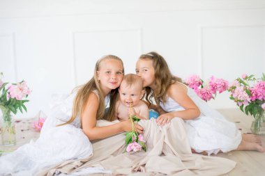 Beyaz elbiseli şirin küçük kız kardeşler ve pembe ve beyaz şakayıklarla süslenmiş açık bahar stüdyosunda bir erkek kardeş.