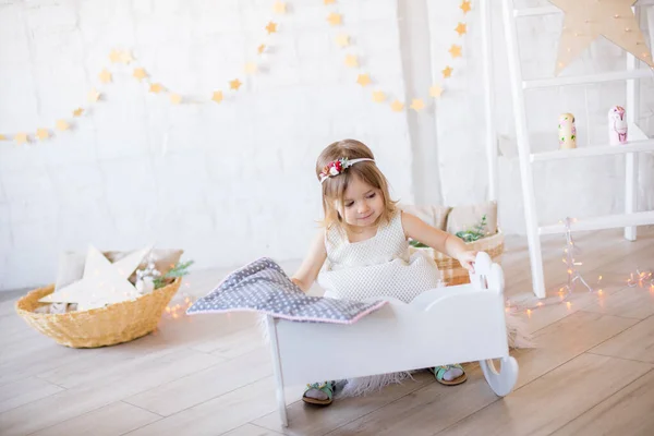 穿着白色连衣裙的可爱小女孩在一间明亮的儿童房的洋娃娃床上玩耍 房间里装饰着玩具和花环 — 图库照片