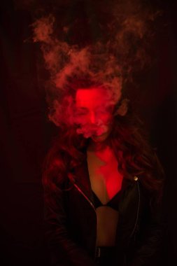 Uzun kıvırcık saçlı genç ve çekici bir kız elektronik sigara içiyor ve kırmızı spot ışıklarına duman salıyor..
