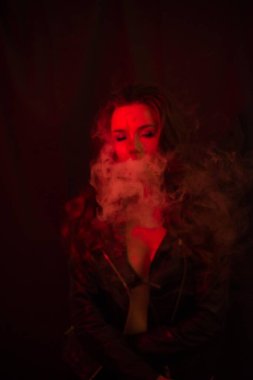 Uzun kıvırcık saçlı genç ve çekici bir kız elektronik sigara içiyor ve kırmızı spot ışıklarına duman salıyor..