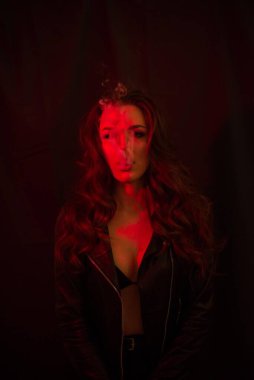 Uzun kıvırcık saçlı genç ve çekici bir kız elektronik sigara içiyor ve kırmızı spot ışıklarına duman salıyor..