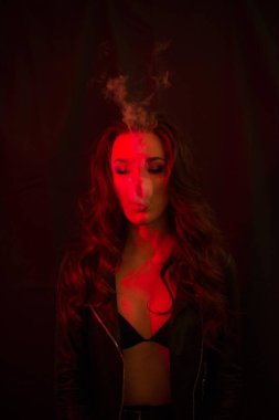 Uzun kıvırcık saçlı genç ve çekici bir kız elektronik sigara içiyor ve kırmızı spot ışıklarına duman salıyor..