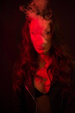 Uzun kıvırcık saçlı genç ve çekici bir kız elektronik sigara içiyor ve kırmızı spot ışıklarına duman salıyor..