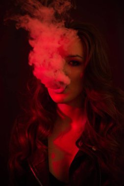 Uzun kıvırcık saçlı genç ve çekici bir kız elektronik sigara içiyor ve kırmızı spot ışıklarına duman salıyor..