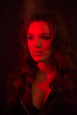 Uzun kıvırcık saçlı genç ve çekici bir kız elektronik sigara içiyor ve kırmızı spot ışıklarına duman salıyor..